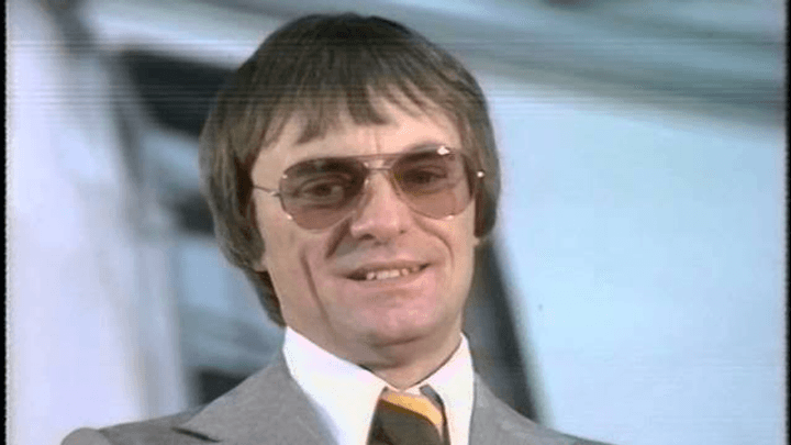 Homme en costume gris et cravate rayée, lunettes de soleil, souriant lors d'un événement