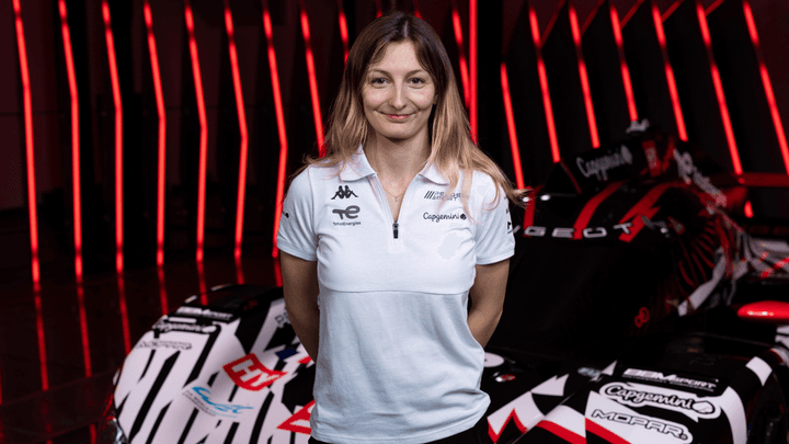 Femme en polo blanc avec logos Kappa et Capgemini devant voitures F1, éclairage rouge
