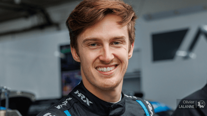 Pilote de Formule 1 souriant en combinaison noire dans le paddock