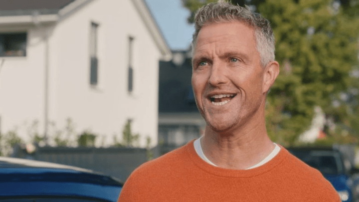 Homme aux cheveux gris en pull orange souriant devant une maison et des voitures