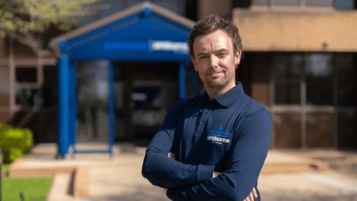 Homme en polo Williams F1 bleu marine, bras croisés, devant bâtiment avec entrée bleue