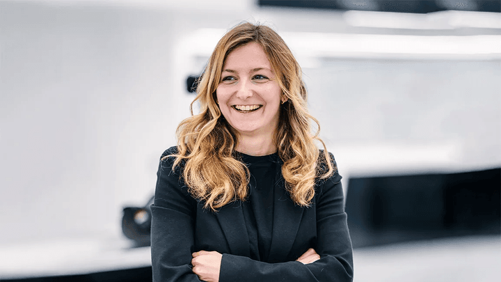 Femme souriante en costume noir, bras croisés, dans un environnement F1 professionnel