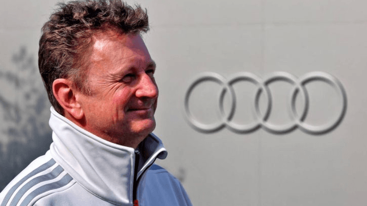Allan McNish en tenue Audi devant le logo de la marque allemande