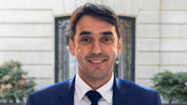 Homme en costume bleu marine et cravate sombre, souriant devant un bâtiment moderne
