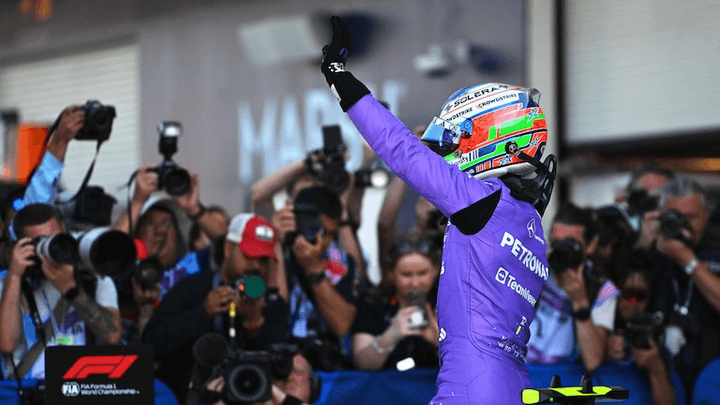 Pilote en combinaison violette célébrant sa pole position, casque aux couleurs vives levé vers le ciel